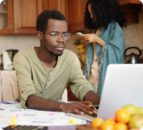 A man using a laptop and a woman who's focused on her phone, standing in the background